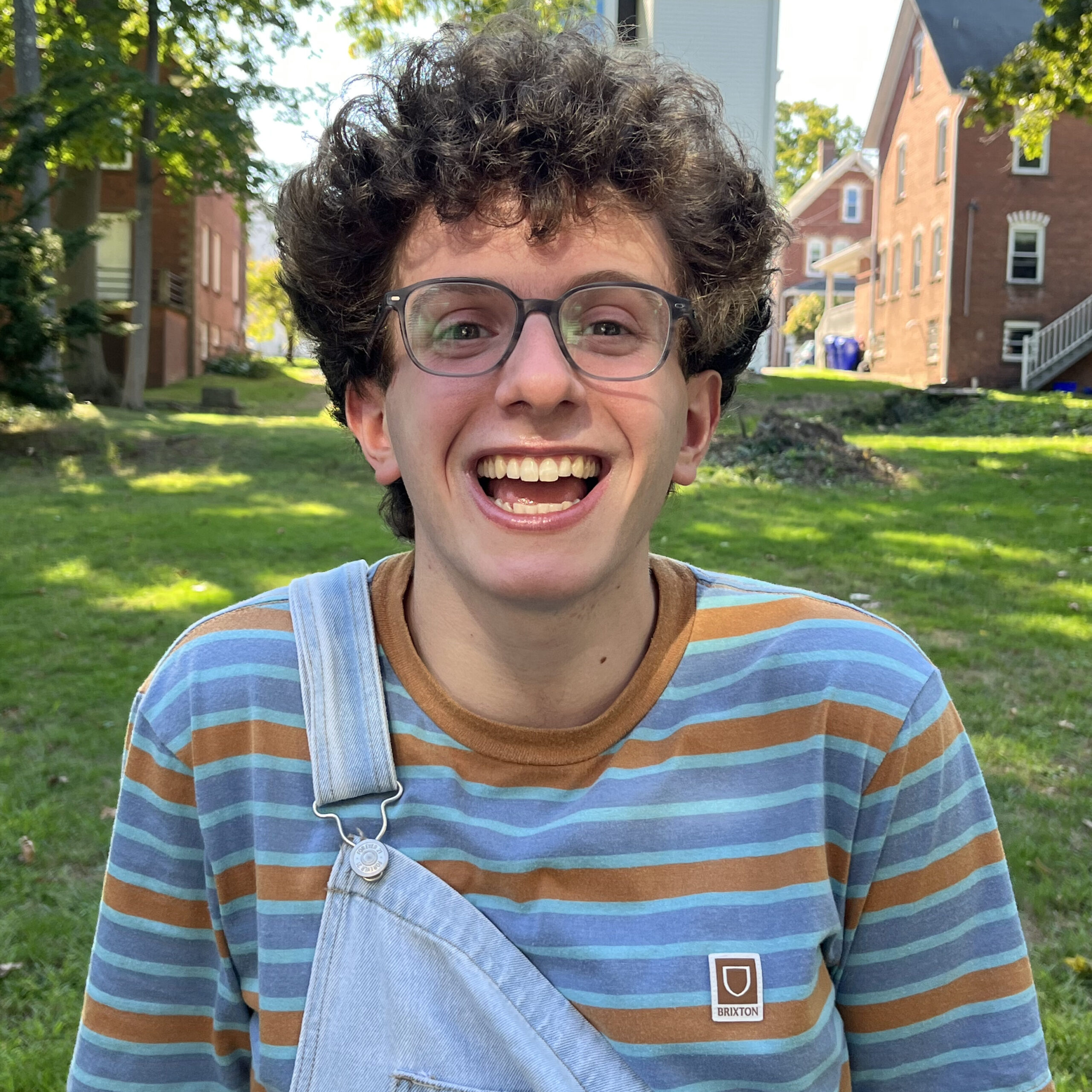 Sam Feuer is standing and smiling in front of a house. He is wearing blue overalls, glasses, and a blue and orange striped shirt.