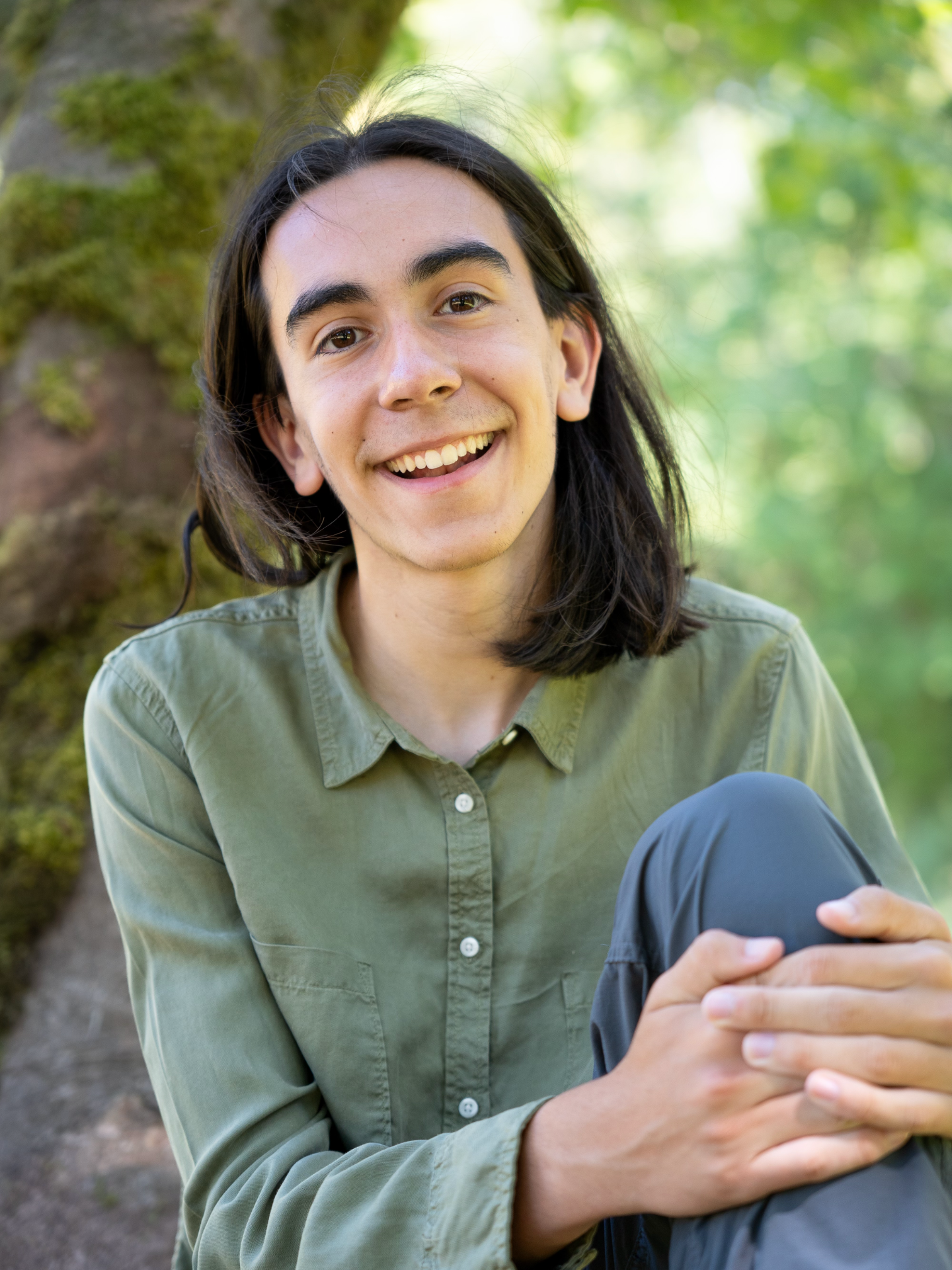 Portrait of Jake sitting on a thick Red Alder tree and clasping their hands together around their left knee.